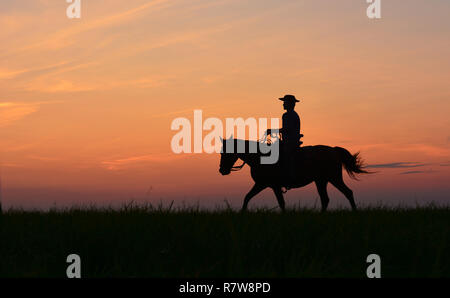 Cowboy in hat riding horse on colorful cloudy sky at sunset. Silhouette of  cowboy travel in wild west mountain like western film background. Stock Photo