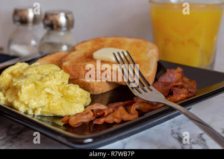 Scrambled eggs, wheat toast with butter, and bacon on square black plate with salt and pepper shakers and orange juice in the background Stock Photo
