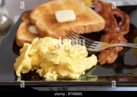 Scrambled eggs, wheat toast with butter and bacon on square plate with fork, up close shot. Stock Photo