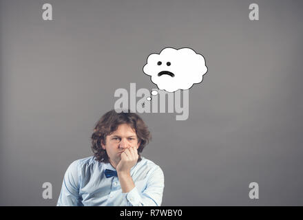 Confused upset handsome young man in blue shirt and bow tie leaned his chin on hand and looking down with drawn cloud of thoughts with sad smile in it Stock Photo