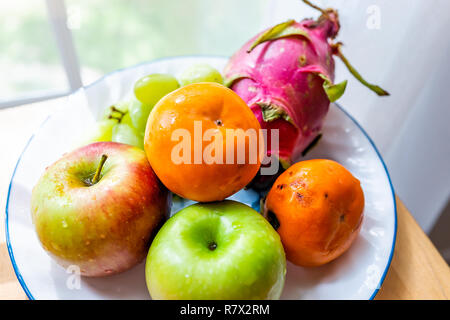 Closeup of whole orange, green, red dragon dragonfruit fruit fruits, apples, persimmons kaki, on plate, wooden table by window, autumn season Stock Photo