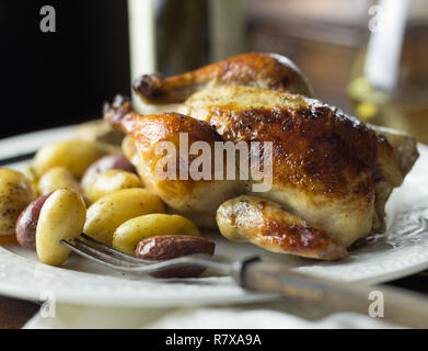 A baked Cornish hen on a plate with a fork, potatoes and white wine. Stock Photo