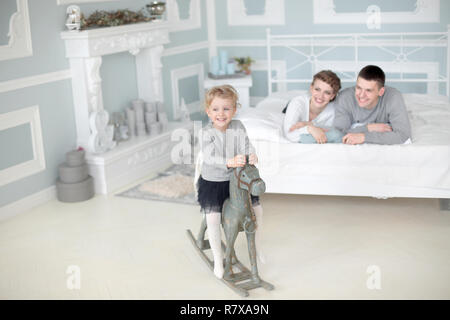 Happy family of three sit in room and play with daughter on her wooden toy horse Stock Photo