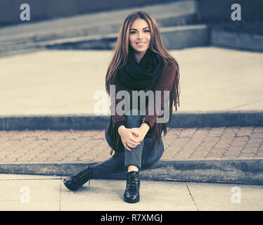 Happy young woman sitting on sidewalk in city street. Stylish fashion model in leather fringe suede jacket and blue jeans Stock Photo