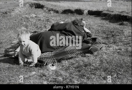 1950s, historical, a woman lying outside on a rug wearing a coat  reading a book with her infant child sitting on the corner,attempting to get its toy, England, UK. Stock Photo