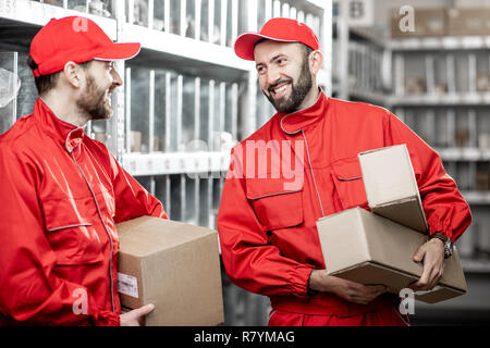 Two handsome warehouse workers in red uniform standing with boxes in the storage with auto parts or other products Stock Photo