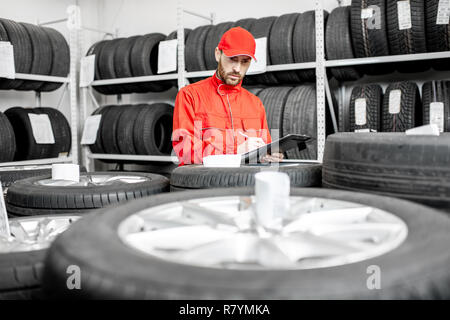 Worker or salesman in red uniform filling some documents checking goods in the warehouse with car tires Stock Photo