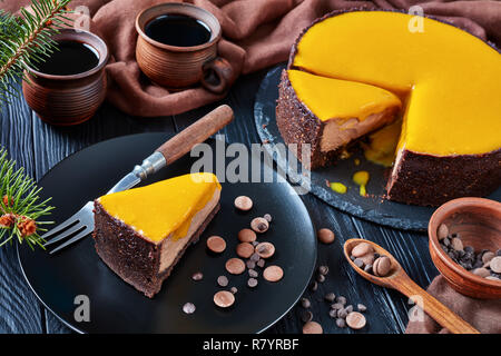 sliced chocolate mango cheesecake. a piece of a cake served on a black plate with dessert fork and chocolate chips. fir tree and cups with coffee on a Stock Photo