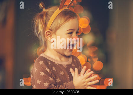 Portrait of a cute little child having fun on kid's Christmas party, baby boy wearing Rudolph horns, laughing and clapping at home near glowing Christ Stock Photo