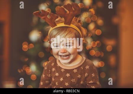 Portrait of a cheerful laughing baby wearing Rudolphs reindeer outfit, with pleasure spending Christmas at home near beautiful glowing Christmas tree Stock Photo