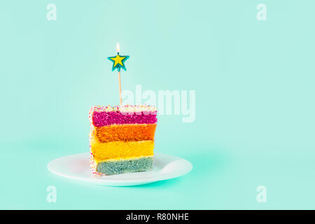 Slice of Rainbow cake with birning candle in the shape of star on white round plate isolated on turquoise background. Happy bithday, party concept. Se Stock Photo