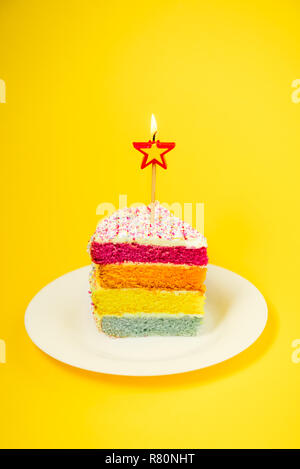 Slice of Rainbow cake with birning candle in the shape of star on white round plate isolated on bright yellow background. Happy bithday, party concept Stock Photo