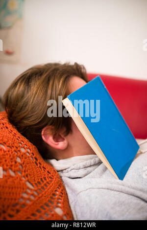 close up side view of a young man lying asleep on a red sofa with a blue hard back cloth bound novel lying across his face Stock Photo