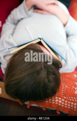 close up aerial view of a young man lying asleep on a red sofa with a blue hard back cloth bound book lying across his face Stock Photo