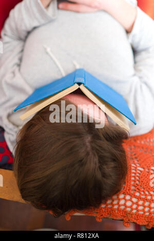 close up aerial view of a young man lying asleep on a red sofa with a blue hard back cloth bound book lying across his face Stock Photo