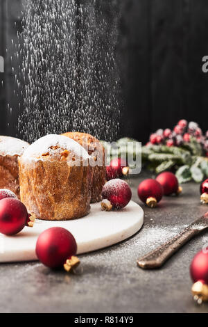 Sprinkling powdered sugar over traditional Christmas mini Panettone with raisins and dried fruits on white marble serving plate surrounded by red baub Stock Photo