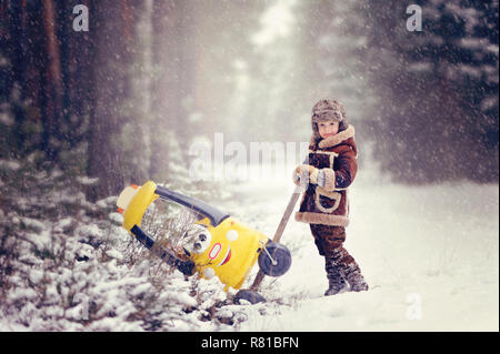 Child playing outdoors during snowfall. The boy pushes from the snow drifts yellow little car, with two teddy bears inside, beautiful snowy winter par Stock Photo