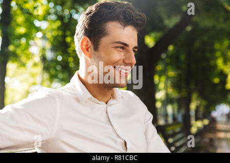 Close up of handsome smiling man dressed in shirt sitting on a bench at the park Stock Photo
