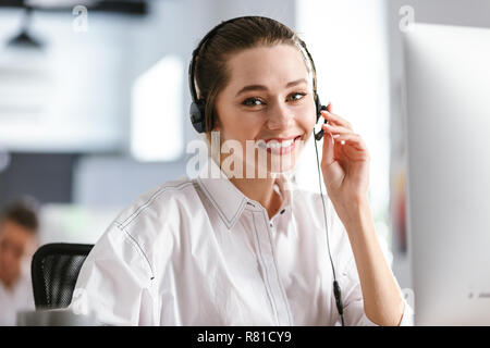 Happy young woman wearing microphone headset dressed in shirt sitting at her workplace at the office, speaking with client Stock Photo