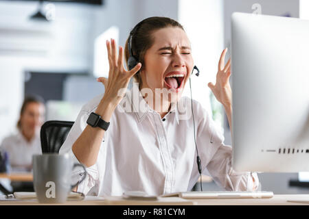 Angry young woman wearing microphone headset dressed in shirt sitting at her workplace at the office, screaming Stock Photo