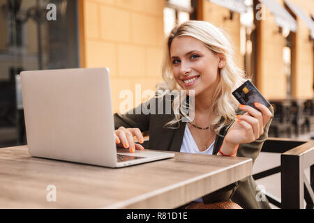 Portrait of adorable blond woman wearing jacket using silver laptop and holding credit card while sitting in cafe outdoor Stock Photo