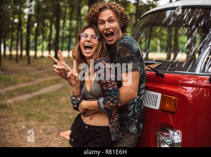 Joyful hippie couple man and woman smiling and hugging each other while standing near minivan in forest Stock Photo