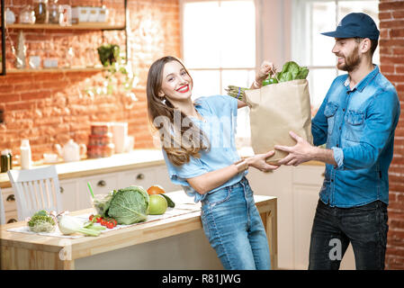 Courier service worker delivering fresh food, giving shopping bag to a happy woman client on the kitchen at home Stock Photo
