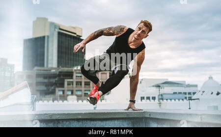Man doing fitness training on rooftop. Muscular man jumping on to the terrace from the rooftop fence with one hand on fence. Stock Photo