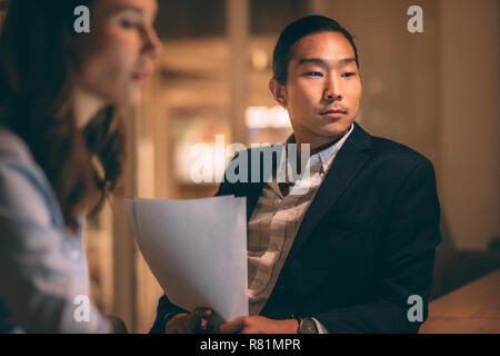 Asian businessman sitting in office holding business papers with his business partner. Blurred close up of a woman working in office with her male col Stock Photo