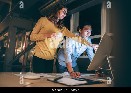 business colleagues discussing business work late in office. Woman entrepreneur pointing at the computer screen explaining to her business partner. Stock Photo