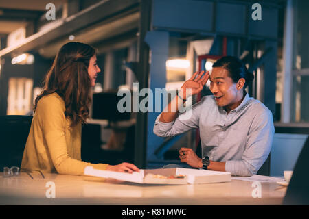 Smiling asian businessman sitting with a female colleague at office eating pizza. Cheerful business partners sitting late in office eating food. Stock Photo