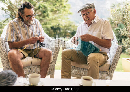 Two senior men sitting on armchair and knitting. Retired male friends doing knitting to pastime at home. Stock Photo