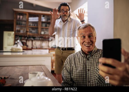 Funny senior man taking selfie with friend standing at back making hand gestures. Retired man making funny face while taking a self portrait with smar Stock Photo