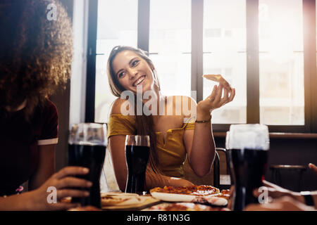 Beautiful young woman sitting at restaurant holding a pizza slice looking at her female friend. Female friends enjoying lunch at cafe. Stock Photo