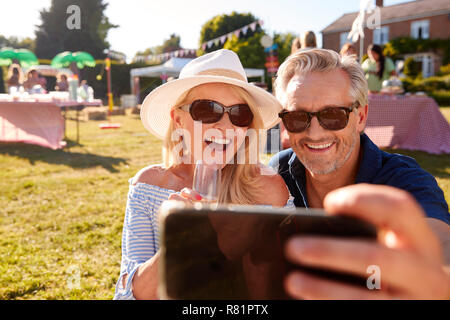 Mature Couple Sitting On Rug At Summer Garden Fete Taking Selfie On Mobile Phone Stock Photo