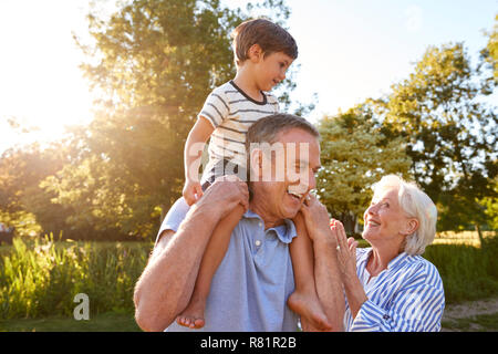 Grandparents Giving Grandson Ride On Shoulders In Summer Park Against Flaring Sun Stock Photo