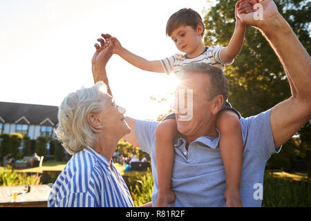Grandparents Giving Grandson Ride On Shoulders In Summer Park Against Flaring Sun Stock Photo