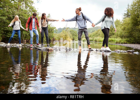 Young adult friends reaching to help each other cross a stream balancing on stones during a hike Stock Photo