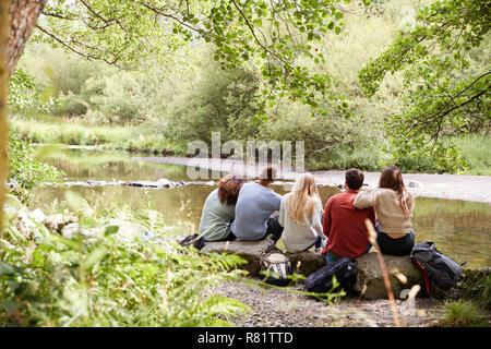 Five young adult friends taking a break sitting on rocks by a stream during a hike, back view Stock Photo