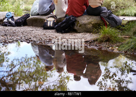 Reflection in stream of five young adult friends taking a break sitting on rocks during a hike, back view, low section Stock Photo