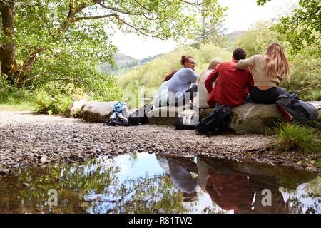 Five young adult friends taking a break sitting on rocks by a stream during a hike, back view Stock Photo