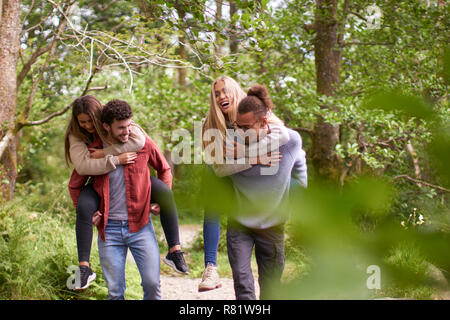 Four smiling young adult friends walking in a forest during a hike, men piggybacking their girlfriends Stock Photo