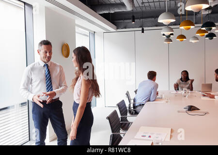 Businessman And Businesswoman Standing In Modern Boardroom Having Informal Discussion With Colleagues Meeting Around Table In Background Stock Photo