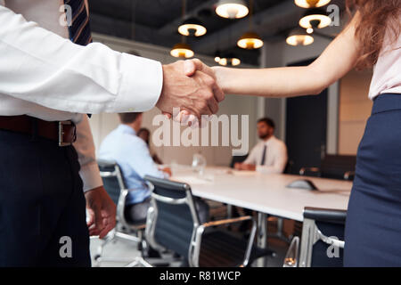 Close Up Of Businessman And Businesswoman Shaking Hands In Modern Boardroom With Colleagues Meeting Around Table In Background Stock Photo