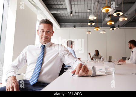 Portrait Of Mature Businessman Sitting In Modern Boardroom With Colleagues Meeting Around Table In Background Stock Photo