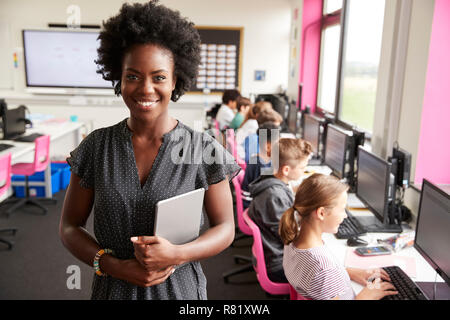Portrait Of Female Teacher Holding Digital Tablet Teaching Line Of High School Students Sitting By Screens In Computer Class Stock Photo