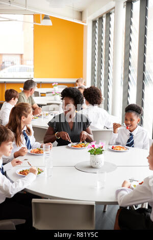 Female Teacher With Group Of High School Students Wearing Uniform Sitting Around Table And Eating Lunch In Cafeteria Stock Photo