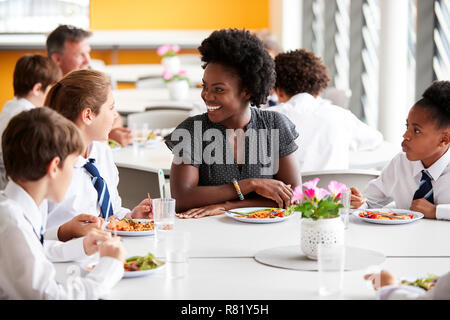 Female Teacher With Group Of High School Students Wearing Uniform Sitting Around Table And Eating Lunch In Cafeteria Stock Photo