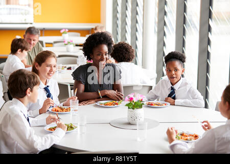 Female Teacher With Group Of High School Students Wearing Uniform Sitting Around Table And Eating Lunch In Cafeteria Stock Photo