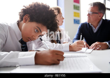 Male High School Student Wearing Uniform Working At Table With Teacher Talking To Pupils In Background Stock Photo